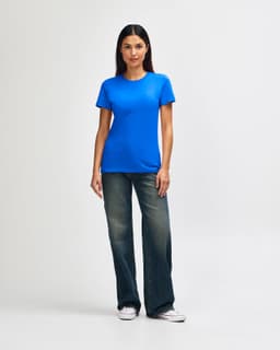 A woman stands wearing a blue Gildan Softstyle ladies' t-shirt and denim pants, paired with white sneakers.