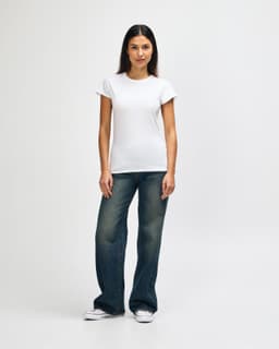 A model wearing a white Gildan Softstyle ladies' T-shirt paired with blue jeans and white sneakers.