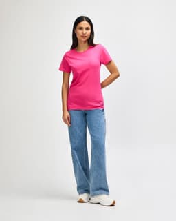 A pink Gildan Softstyle ladies midweight t-shirt, worn with blue jeans and white sneakers. The model stands against a plain background.