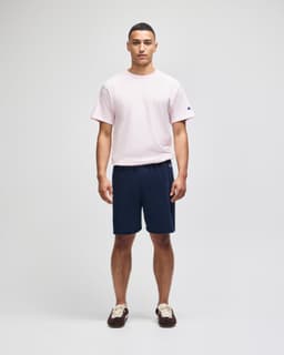 A man stands wearing a pink t-shirt and navy cotton gym shorts, featuring a logo on the shorts.