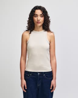 A fitted tank top in a light beige colour, featuring a high neckline and without sleeves. The model is wearing jeans.