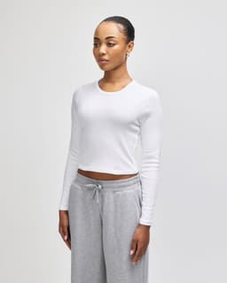 A white long-sleeve crop fitted T-shirt, modelled by a woman, paired with grey sweatpants. The shirt features a logo.