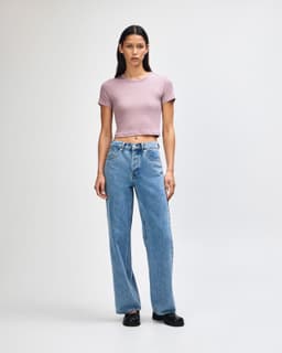 A fitted women's crop T-shirt in soft pink, paired with high-waisted blue jeans. The model stands against a light background.
