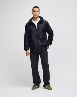 A black full-zip anorak jacket stands on a model, styled with a white shirt and dark pants. The jacket features a logo.