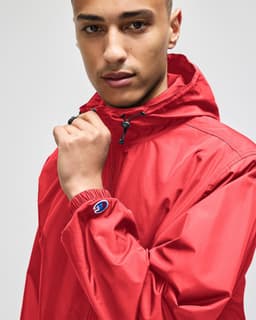 A red full-zip anorak jacket with a hood and an elastic cuff, featuring a logo on the sleeve.