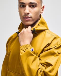 A packable anorak jacket in mustard yellow, featuring a hood and an adjustable neckline. It has a logo on the sleeve.
