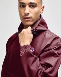 A maroon packable anorak jacket featuring a hood and adjustable cuffs, with a logo on the sleeve.