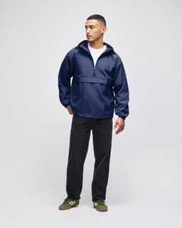 A model wearing a navy blue packable anorak jacket, paired with black trousers and green shoes. The jacket has a logo.