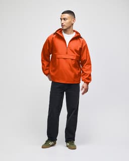 An orange packable anorak jacket worn by a man, paired with dark trousers and sneakers, featuring a hood.