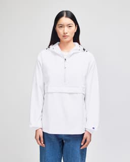 A white packable anorak jacket with a hood and a front pocket, featuring a logo. Model is wearing blue jeans.