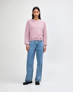 A light pink women's crewneck sweatshirt paired with blue wide-leg jeans and white sneakers. The sweatshirt has a logo.