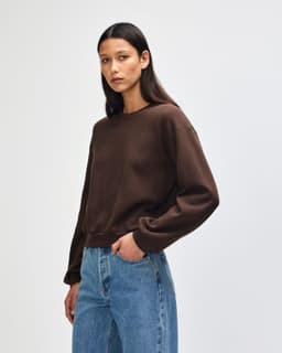 A brown women's crewneck sweatshirt with long sleeves, featuring a fitted hemline. The model is wearing denim jeans.
