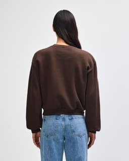 A women's crewneck sweatshirt in brown, featuring a relaxed fit and a logo. The model is shown from the back.