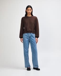 A woman stands confidently in a brown crewneck sweatshirt and light blue jeans, wearing white sneakers.