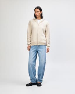 A unisex full zip hooded sweatshirt in beige, worn by a model standing in black cargo pants and sneakers.