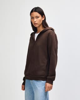 A unisex full zip hooded sweatshirt in brown, featuring a front zipper and a hood. The model stands with hands in pockets.