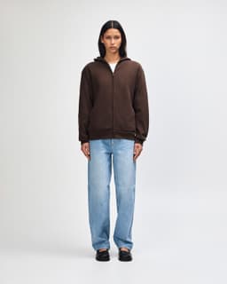 A unisex full zip hooded sweatshirt in brown, worn with light blue baggy jeans and sneakers. Features a logo.