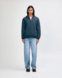A unisex full zip hooded sweatshirt in navy, worn with baggy light blue jeans. Features a logo on the front.