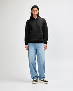 A unisex black hooded sweatshirt is worn with light blue baggy jeans and black sneakers, featuring a logo.