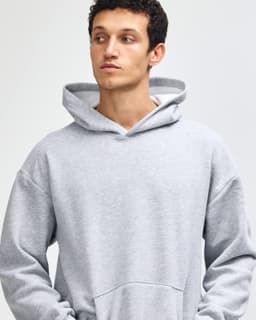A grey unisex hooded sweatshirt with a front pocket and a logo. The model is standing with hands in pockets.