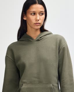 A unisex hooded sweatshirt in olive green with a front pocket and a logo. The model is wearing it casually.