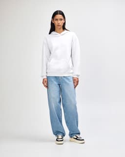 A unisex hooded sweatshirt in white, featuring a front pocket and a plain design. The model wears casual light blue jeans.