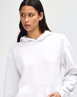 A unisex hooded sweatshirt in white, featuring a pocket and a logo on the front. The model adjusts the collar.