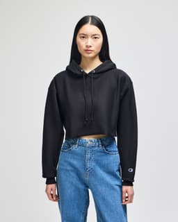 A black cropped hoodie with a hood and long sleeves, worn with blue jeans. It features a logo on one sleeve.