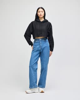 A woman wearing a black cropped hoodie with loose blue jeans and white sneakers, standing against a plain background.