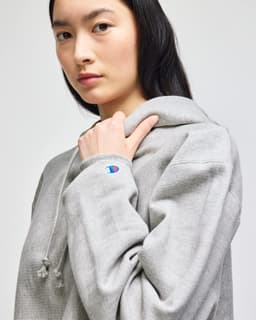 A grey cropped hoodie with a large hood and a logo on the sleeve, modelled by a woman.