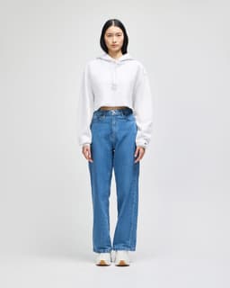 A cropped white hoodie with a drawstring hood paired with blue jeans, modelled by a woman. The hoodie features a logo.