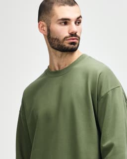 A model wearing an olive green Gildan Softstyle adult sweatshirt. The sweatshirt has a round neckline and long sleeves.