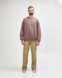 A male model wears a dusty pink Gildan Softstyle Adult Sweatshirt with black pants and black sneakers. The sweatshirt has a simple design.