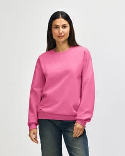 A pink Gildan Softstyle adult sweatshirt is worn by a model, paired with blue jeans. The sweatshirt has a logo.