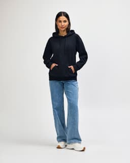 A black Gildan Softstyle adult hooded sweatshirt worn by a model with dark jeans and sneakers, featuring a logo.