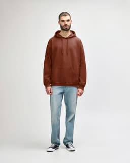 A woman wearing a brown Gildan Softstyle Adult Hooded Sweatshirt and blue jeans, standing against a plain background.