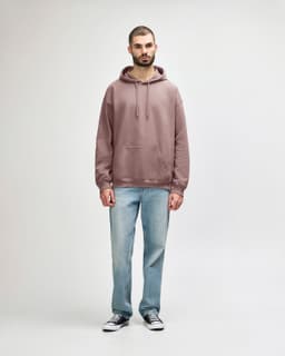A woman wearing a dusty rose hooded sweatshirt paired with blue jeans and white shoes. The sweatshirt has a logo.