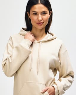 A model wearing a beige Gildan Softstyle adult hooded sweatshirt with the hoodie drawn over her head.