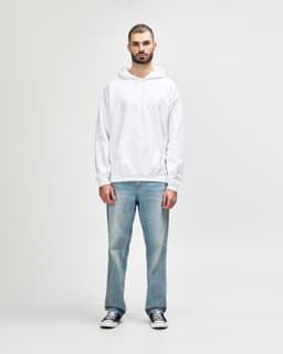 A man stands in a white hooded sweatshirt and light blue jeans, wearing black shoes. The sweatshirt has a logo.