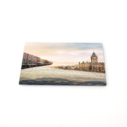 A custom tinplate fridge magnet featuring a scenic view of a river and buildings, with soft pastel colours.