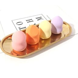A tray holds four gourd-shaped makeup sponges in pink, orange, yellow, and purple. The tray is gold in colour.