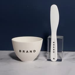A white mixing bowl and spatula, both featuring a logo, arranged on a marble surface against a dark background.