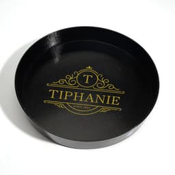 A round, black non-slip tray featuring a decorative logo in gold.