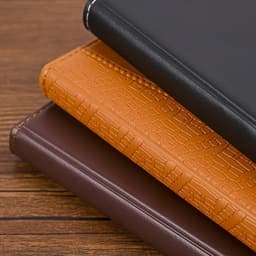 Three diaries stacked: one black, one orange with a textured pattern, and one brown, all featuring a subtle logo.