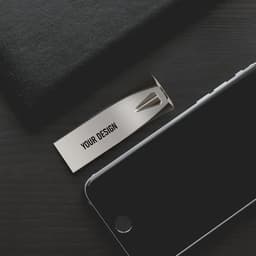 A silver 32 GB USB flash drive rests on a dark wooden surface next to a black smartphone and a black notebook.