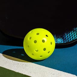 A bright yellow pickleball with holes sits on a blue and white surface next to a paddle with a textured design.