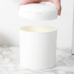A white mini desktop trash can with a removable lid, sitting on a marble surface.