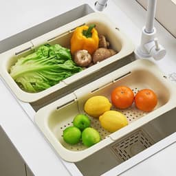 An extendable kitchen sink colander in cream, featuring lettuce, mushrooms, a bell pepper, and assorted fruits.