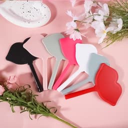 A collection of lip-shaped hand mirrors in black, grey, pink, white, and red, arranged on a pink background with flowers.