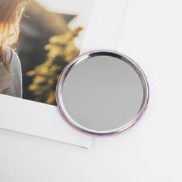 A circular tinplate pocket mirror with a silver reflective surface and a pink border, placed next to a photograph.